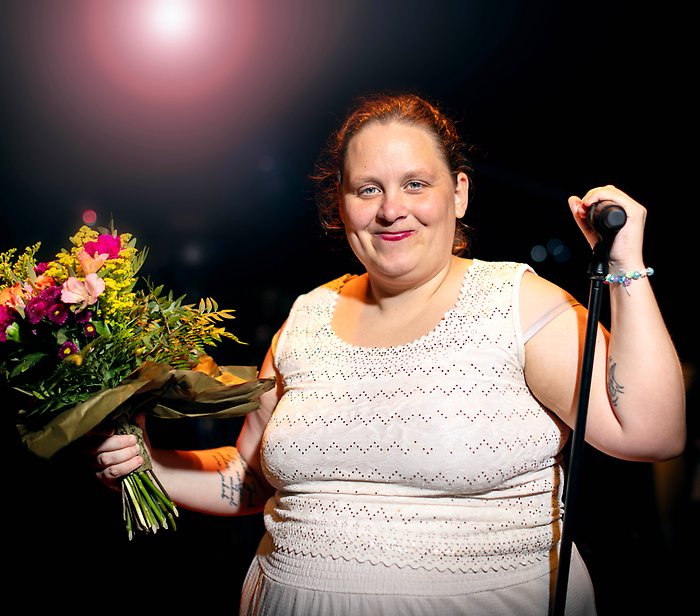 Bild på Nikki. Hon har en vit klänning på sig och ler. Hon står på en scen och håller upp en blombukett.