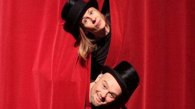 En kvinna och en man tittar fram med huvudena genom glipan i en röd ridå. Båda har hög hatt,.