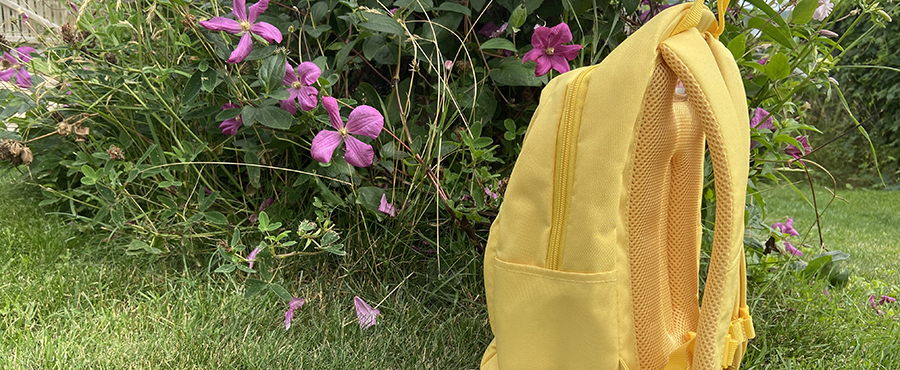 En gul ryggsäck och i bakgrunden syns lila blommor och en grön gräsmatta. 