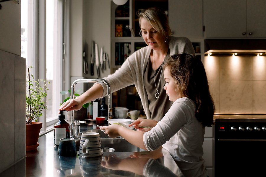 Mor och dotter i kök vid diskbänk med rinnande vattenkran.