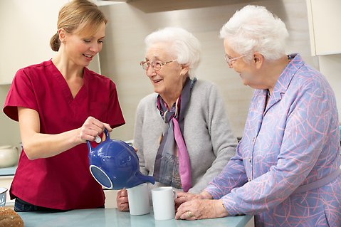 En ung kvinna inom omsorgen häller upp varm dryck i koppar till två äldre damer.