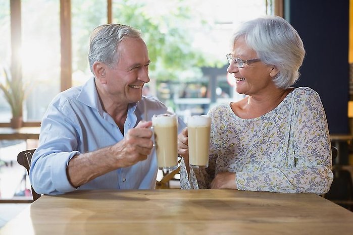En äldre man och kvinna tittar på varandra och ler. I en hand har de varsitt högt glas med kaffe och mjölk.