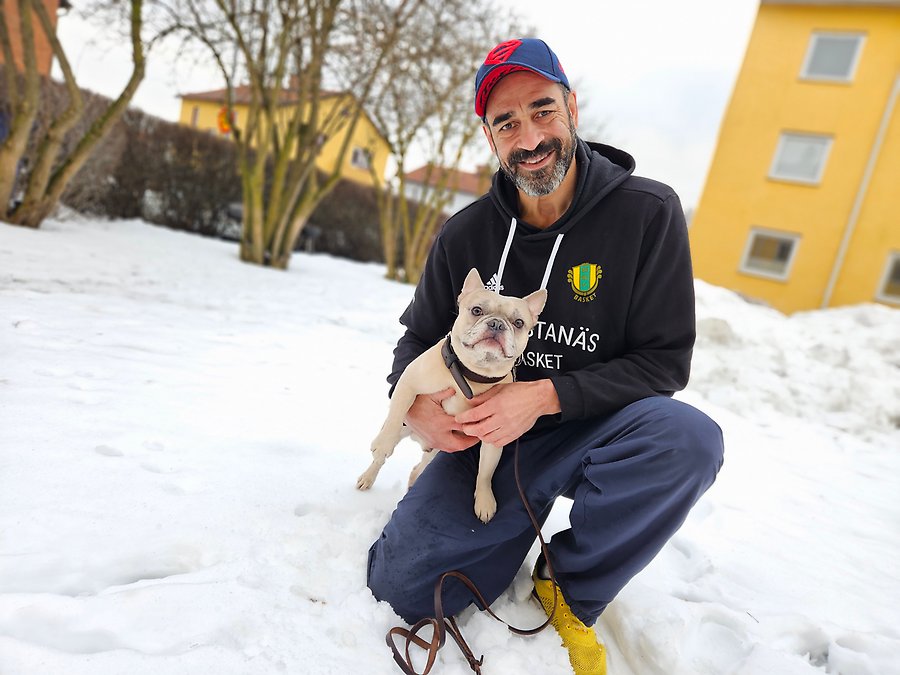 En man sitter på huk bredvid sin hund och ler in i kameran.