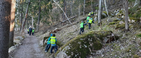 Förskolebarn med gula västar klättrar upp för ett berg. I bakgrunden syns en pedagog och några fler barn i gula västar.