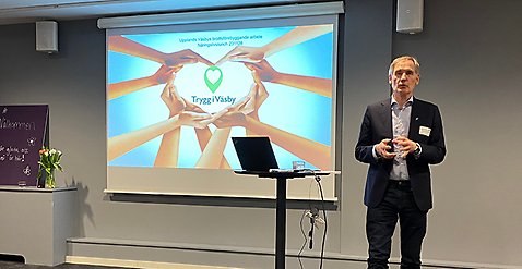 En man står och presenterar. Han står vid ett ståbord med en dator och i bakgrunden syns en bild på händer som formar ett hjärta. I mitten av hjärtat syns en text, Trygg i Väsby.