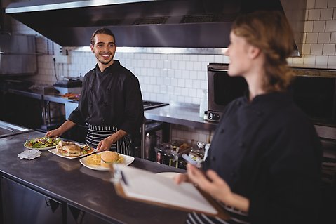 En unga man står bakom en bänk i ett restaurangkök, några tallrikar med mat är placerade framför honom. En ung kvinna står på andra sidan bänken och väntar på att leverera beställningen.