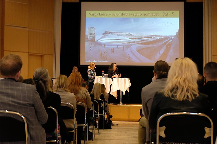Lena Nordenlöw, stadsarkitekt och Åsa Engwall, chef för kontoret för samhällsbyggnad står på en scen och presenterar stadsbyggnadsprojektet Väsby Entré som syns på en stor skärm.