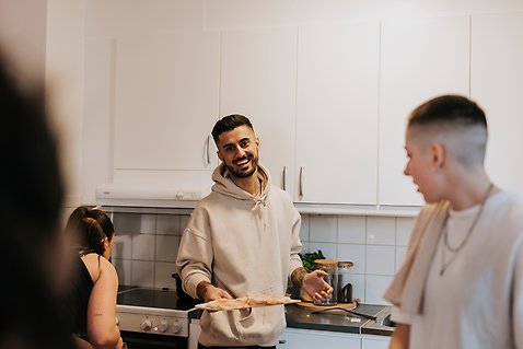 Tre unga personer står i ett kök och pratar med varandra medan de lagar pizza. 