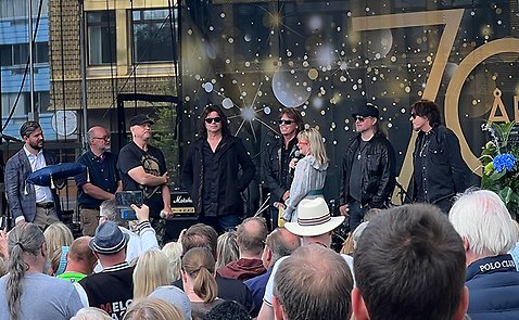 På en scen står alla fem medlemmar från bandet Europe, tillsammans med styrelseordförande Oskar Weinmar, konferencier Josefin Sundström och Roland Storm. Nedanför scenen syns en publik. 